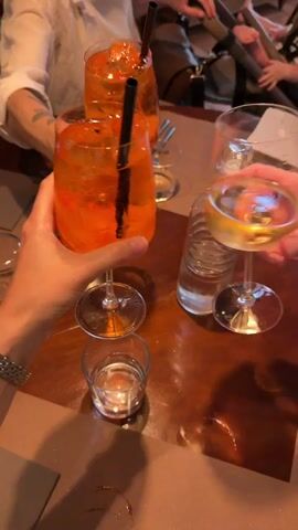 The video opens with a close-up shot of hands holding glasses of drinks. In the foreground, a hand with a silver bracelet on its wrist is holding a tall glass filled with an orange-colored beverage and ice, with a black straw. To its right, another hand is holding a wine glass with a pale yellow liquid and ice. Behind these, two more tall glasses of the same orange beverage are visible, also with straws. The drinks appear to be on a wooden table. In the background, out of focus, parts of people are visible, suggesting a social gathering. The overall impression is a toast or a celebration.