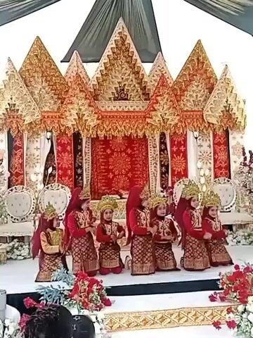 A group of young children in traditional attire are performing a dance on a stage. The stage is decorated with elaborate, golden and red structures resembling traditional houses, with draped fabrics and floral arrangements. The children are dressed in matching red and gold outfits, complete with ornate headwear. They are arranged in two rows, with some of them kneeling and others standing, performing synchronized movements. In the foreground, two young girls are visible, facing the stage and appearing to watch the performance. The overall setting suggests a cultural event or celebration.