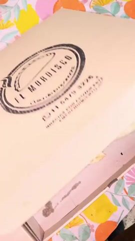 Here is a description of the video:

A camera pans downward into an open cardboard box containing an assortment of baked goods. The box rests on a surface with a colorful citrus-themed tablecloth.  Most of the pastries are folded into a crescent shape, and some have a crimped edge. There are also several open-faced pastries, topped with various fillings that include corn, white cheese, tomatoes, and black olives. The pastries are slightly shiny, indicating they may have been brushed with oil or egg wash before baking. The inside of the box has a light-colored paper liner. Background audio plays music and talking.