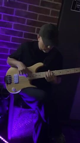 The video shows a man playing a light-colored bass guitar. He is wearing a black t-shirt and a dark baseball cap that obscures his face. He is standing near a brick wall with purple lighting. His left hand is on the neck of the guitar, while his right hand is strumming. In the background, another musician is visible, and a chalkboard displays what appears to be a set list or schedule. The lighting is dim, with a strong purple hue.