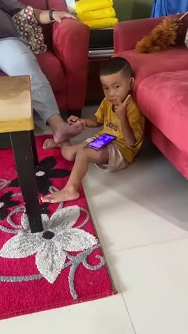 A young boy in a yellow t-shirt and khaki shorts sits on the floor next to a red sofa, holding a smartphone. He is looking towards the camera and holding up two fingers. His left foot is extended towards a wooden table. A person's legs, wearing gray pants, are visible on the left side of the frame, seated on the red sofa. The floor is covered by a red rug with a floral pattern. In the background, there is another red sofa, yellow pillows, and a duster. The scene appears to be indoors, likely a living room.