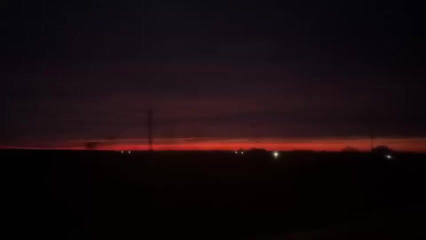 The video shows a dark, pre-dawn or dusk landscape. A dark horizon line is visible against a sky that is a gradient of deep reds and purples. Sparse, indistinct shapes that may be trees or bushes are silhouetted against the faint light. Small, scattered pinpoints of light, possibly from distant buildings or vehicles, dot the horizon. A tall pole-like structure is also visible on the horizon, appearing as a silhouette. There's a sense of movement in the video, likely from a moving vehicle, causing the lights to shift slightly.