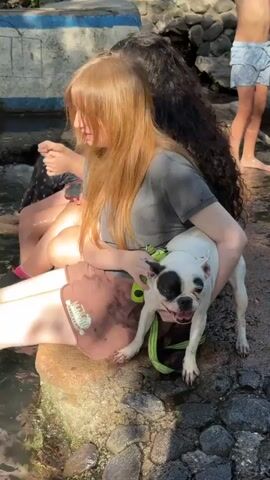 A young woman with long, reddish-blonde hair is sitting on a rock by a body of water, holding a white French bulldog with black markings. The dog is wearing a bright green harness. The woman is wearing a dark gray t-shirt and brown shorts. To her left, another person with dark, curly hair is also sitting on the rock. In the background, a young person in blue swim trunks stands in the water, and a yellow inflatable ring is visible. The environment appears to be outdoors with rocks and vegetation.