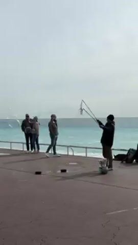 The scene shows a seaside promenade. A man in a dark jacket and beanie is using a large, multi-loop bubble wand to create giant soap bubbles. Several people are watching the bubbles. Two are standing close to the railing, and another is standing a short distance away. The railing is metal and runs along the edge of the promenade, separating it from the turquoise sea. The sky is overcast. Some black rectangular objects are scattered on the paved area, near a bucket.