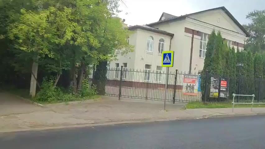 The video shows a building with a white facade and a mansard roof, surrounded by a black metal fence and a row of tall, green trees. A pedestrian crossing sign is visible in front of the building. The building appears to be an institutional or cultural establishment, possibly a museum or an educational institution, based on the posters displayed on the fence. The posters are in Russian and feature images of military personnel, suggesting an exhibition related to war or military history. The environment is urban, with a road and sidewalk in the foreground. The weather is overcast, creating a muted lighting effect. The video appears to be taken from a moving vehicle, as indicated by the slight blur and motion perspective.