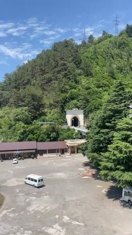 The video pans from a tunnel entrance on a forested mountainside down to a parking lot with a few parked vehicles, including a white van and a white car. There are also several small buildings with awnings. Further in the distance, a bridge crosses over a road where cars are driving. Beyond the bridge, there are more buildings, trees, and a glimpse of the sea. The sky is blue with some white clouds. The camera movement is smooth, showing a panoramic view of the area.