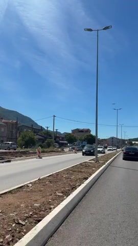 This video shows a clear blue sky with a few wispy clouds above a street scene. The road appears to be under construction or renovation, with bare earth and some construction barriers visible on the left side. Several cars are driving on the road. In the background, there are buildings and a hill or mountain. The video is taken from a moving vehicle, as indicated by the slight blur and the perspective.