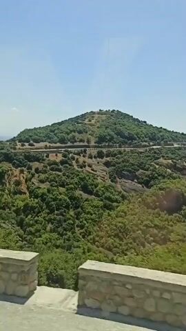The video shows a view of a mountainous landscape with a clear blue sky. Lush green trees cover the hillsides, and some of the terrain appears rugged and rocky. In the foreground, there are stone walls that seem to line a road. The camera is moving, giving the impression of a journey through this scenic environment. There are no people visible in the video. The overall impression is one of natural beauty and a sense of exploration.