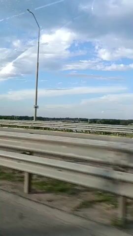 A camera pans across a road, with a blurry car interior in the foreground. In the background, a highway with multiple lanes is visible, separated by guardrails. Beyond the highway, there are trees and a body of water, likely a river or lake, under a cloudy blue sky. A tall street lamp is also visible on the right side of the frame. The video appears to be shot from a moving vehicle.