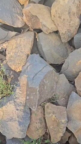 The video shows a close-up, overhead view of a pile of rocks. The rocks are of various sizes and shapes, with some being angular and others more rounded. The colors of the rocks range from light brown and tan to dark gray and black. There are small patches of green vegetation growing between some of the rocks. The lighting suggests it is daytime, with sunlight casting shadows on the rocks. No people or actions are visible.