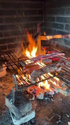 This video shows a close-up, low-angle shot of a barbecue grill. The grill is made of metal grates suspended over a bed of glowing red embers and active flames. Several types of meat are cooking on the grill, including what appear to be sausages and cuts of steak. One sausage is being held over the grill by a pair of tongs. Sparks are flying from the fire. The grill is situated within a brick structure, likely a fireplace or outdoor cooking area. The lighting is dim, with the fire providing the primary light source.