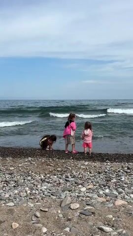 The video shows three young children on a rocky beach next to a large body of water. The child on the far left is bending down, appearing to be playing with pebbles on the shore. The child in the middle is standing and facing the water, wearing a pink jacket and a patterned skirt. The child on the right is also standing and facing the water, wearing a pink hooded jacket and shorts. Waves are breaking on the shore, indicating that the water is active. The sky is blue with some scattered clouds.