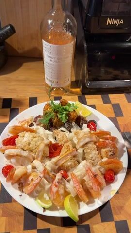 A plate of food is centered in the frame. The dish appears to be pasta with shrimp, tomatoes, and some sort of breaded topping. There are also pieces of lime on the plate. Behind the plate, a bottle of rosé wine is visible, with a black appliance, possibly a blender, in the background. The scene is set on a wooden surface. The video is a close-up, static shot of the prepared meal.