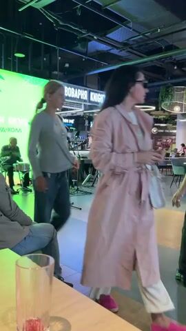 The video opens with a wide shot of an indoor public space, likely a food court or entertainment venue, with a live band performing on a raised stage to the left. A young blonde girl in a grey long-sleeved shirt and blue jeans walks past the camera towards the center of the frame. In the background, a woman in a patterned dress stands near the stage, and other band members are visible.

As the video progresses, the focus shifts to the right side of the frame. The blonde girl continues to walk, and a man in a black t-shirt and black pants is seen dancing. A little girl in a white t-shirt and a bright orange tutu is also dancing, twirling near a man in a green jacket and a cap. Another man in a green shirt is seated on a green couch in the background. The environment features modern lighting fixtures, hanging plants, and various seating areas. The camera pans and zooms slightly, capturing the interactions and activities within the space. The band continues to play music, and people are moving about and enjoying themselves.
