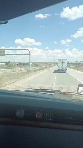 A view from inside a vehicle, looking out the windshield at a highway. A white semi-trailer truck with the word "CRST" on the back is driving on the highway ahead. The sky is blue with white clouds. The interior of the vehicle shows a dashboard with some objects on it, including a book or stack of papers. The road is paved, and there is a shoulder on the right side of the highway. There are some buildings and fences visible in the distance. The video is short and appears to be a snapshot of a drive.