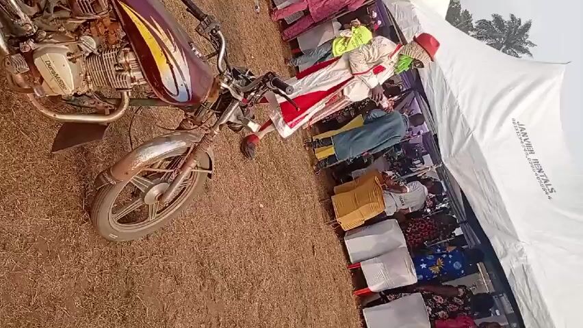 The video depicts a man dressed in a white and red ceremonial outfit with a red hat, walking in front of a seated audience under a white tent. The man is the central figure, and he is walking with purpose, possibly performing a dance or ritual. The audience is seated on white chairs. To the left of the scene is a motorcycle. The ground is covered in dry grass. The tent's white material dominates the top portion of the frame.