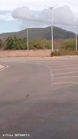 The camera pans across a deserted parking lot surrounded by green foliage, mountains, and a cloudy sky.  A man with a backpack featuring the colors of the Venezuelan flag walks across the parking lot, followed by a man on a bicycle. The scene is quiet and rural.