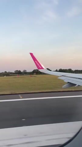 The video shows the view from an airplane window during takeoff or landing. The right wing of the plane is prominently visible, featuring a pink tip with the Wizz Air logo. Below the wing, a runway is seen, marked with white lines. Beyond the runway, a grassy field stretches out, bordered by trees and a soft pink and blue sky. The sun appears to be low on the horizon, suggesting either dawn or dusk. The overall impression is of a peaceful ascent or descent during a beautiful time of day.
