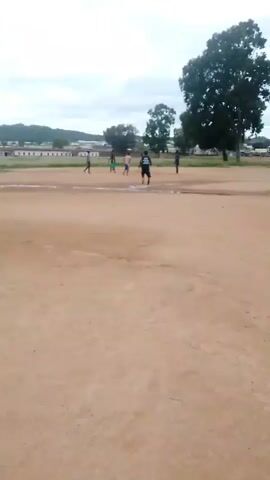 A group of five boys are playing soccer on a dirt field. The sky is overcast. In the background, there are buildings and trees.
