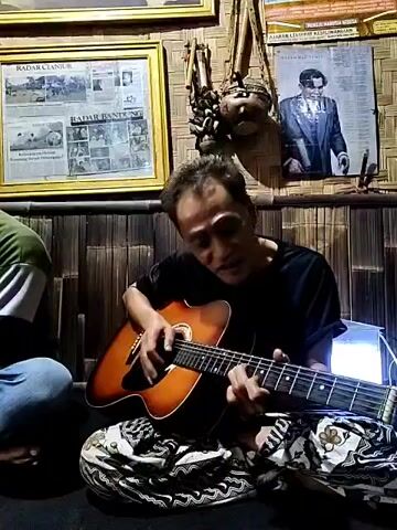 A man sits cross-legged on a cushion, enthusiastically playing an acoustic guitar and singing. He wears a black t-shirt and patterned pants. Another person sits to his left, wearing a green shirt and blue jeans, partially visible in the frame. The backdrop includes a bamboo-like wall adorned with framed newspaper clippings, a picture of a man in a suit, and what appears to be traditional Indonesian artifacts. The environment suggests an intimate, possibly home-based setting.