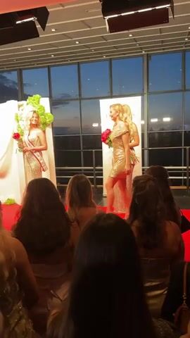 The scene captures a pageant event where a young woman in a red dress and crown is on stage, receiving or giving a sash to another woman in a gold dress.  Surrounding them are two other women in similar formal attire, sashes, and carrying bouquets. All the women are standing on a red carpet in front of a backdrop decorated with white fabric, floral arrangements, and a branded sign. The audience, comprised of people of varying ages and hairstyles, are observing the scene, some holding up cell phones to take pictures or videos.  The event appears to be taking place in a ballroom with large windows providing a view of the evening sky. The overhead lighting casts a warm glow on the stage.