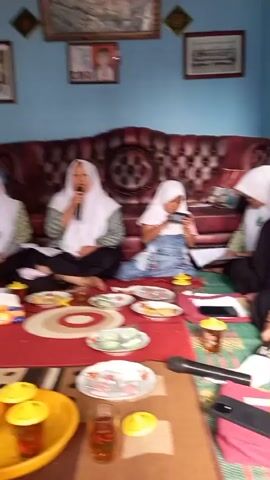 The video shows a group of five young women, all wearing white hijabs and patterned long-sleeved tops, sitting on the floor in a room. Three of the women are facing the camera and appear to be engaged in conversation, with one woman holding a microphone and speaking. The other two women are looking down, possibly at books or papers. In the foreground, there are several plates of food, glasses of tea, and a microphone on a red mat. The room has blue walls and a large red sofa. The lighting is dim.