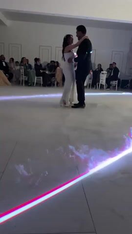 A newly married couple dances on a cloud-covered dance floor. The bride is wearing a long, white strapless dress and the groom is wearing a dark suit. Guests sit at tables surrounding the dance floor. A light emitting a pink and blue spectrum shines on the front of the dance floor.