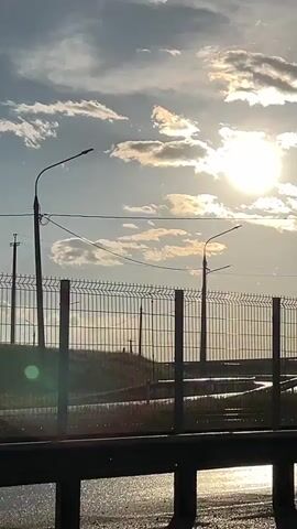 The video shows a view from a moving vehicle on a road. In the foreground, a dark barrier is visible. Beyond the barrier is a chain-link fence with a grassy embankment behind it. Two streetlights are visible, one on the left and one on the right, with power lines connecting them. The sun is shining brightly in the sky, partially obscured by clouds, creating lens flares. In the middle of the frame, a road sign with the designation "M4" is visible on a pole. The overall scene depicts a roadside environment during daylight hours.