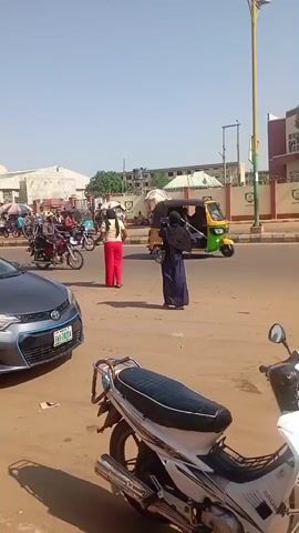 The video shows a street scene with several people and vehicles. In the foreground, a motorcycle is parked. Further back, a dark gray sedan is parked on the left side of the road, with its front bumper visible. To the right of the sedan, two individuals are standing on the street. One person is wearing a dark headscarf and a long blue garment, while the other is wearing a white top and red trousers. Behind them, several other people are visible, along with motorcycles and a tricycle. In the background, there are buildings and streetlights. The road appears to be paved, and the ground on the side of the road is dirt or gravel. The sky is clear and blue. There are utility poles and wires visible along the street. In one segment, a young boy is shown walking on the side of the road carrying a black plastic bag. Another segment shows a red sedan driving on the road.