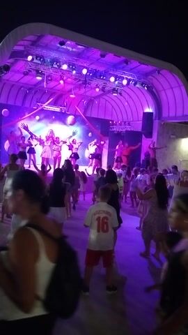 This media file depicts a group of people enjoying an outdoor performance or event at night. The scene is lit with vibrant purple and pink stage lights, creating a festive and energetic atmosphere. A group of performers are on a stage with a cosmic-themed backdrop featuring planets and nebulae. They are illuminated by spotlights and appear to be dancing or engaged in some form of choreographed movement.

In front of the stage, a crowd of spectators are gathered, facing the performers. They are of various ages and appear to be watching the show with interest. The image is taken at an angle, suggesting the viewer is part of the crowd. Some individuals in the crowd are blurred, indicating movement or a lack of focus on them.

Notable details include the presence of stage lighting equipment, speakers, and a large, curved white canopy structure overhead, suggesting a dedicated event space. The attire of the people, including dresses and casual wear, is consistent with a warm evening. The overall mood appears to be one of enjoyment and entertainment. There is no visible indication of the time of day beyond it being night, nor any weather cues. The location context of Caorle, Italy, suggests this could be a summer event at a tourist destination.

A piece of visible text is present on the back of a white t-shirt worn by a person in the crowd. The text reads "16" and below it, "BLASZCZYKOWSKI".