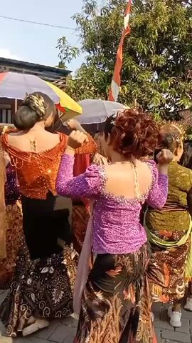 Two women, viewed from behind, are dancing. The woman on the left wears a copper-colored, sequined, off-the-shoulder top with puffed sleeves and a black skirt with a patterned hem. Her hair is pulled back in a bun. The woman on the right wears a bright pink, sequined, off-the-shoulder top with puffed sleeves and a dark patterned skirt. Her hair is styled in an updo. They are in an outdoor area with trees and foliage visible in the background, along with tents or canopies and a flag with red and white stripes. Other people are partially visible in the background.