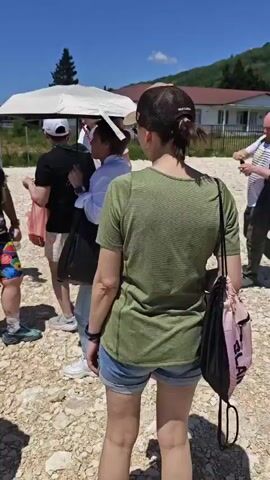 A group of people stands outdoors on a sunny day. The ground is covered with light-colored gravel. A woman in a white shirt and blue jeans holds a large white umbrella over her head and a black bag in her left hand. Next to her, a man in a white hat and white shirt takes something from a plastic bag held by another person. A man in a colorful vest and shorts holds a phone to his ear. Several other people stand around, some wearing hats or caps. In the background, there are houses with orange roofs and green trees under a clear blue sky.