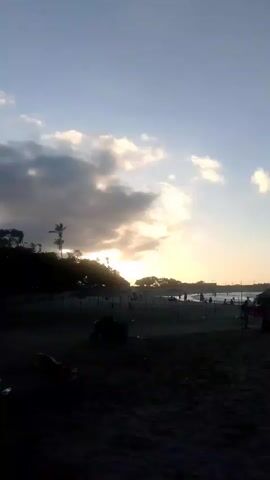 The video shows a beach scene at dusk. The sun is setting behind clouds, casting a warm glow on the sky. There are people on the beach, some sitting in chairs and others walking around. There are also some palm trees and thatched umbrellas visible. The overall atmosphere is relaxed and peaceful.