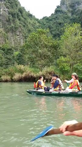 A medium shot shows four people traveling by boat on a waterway with lush, verdant banks. Three of the passengers, wearing life vests, are seated facing the camera and engaged in conversation or snacking, while the fourth, donning a traditional conical hat, stands at the stern, propelling the boat with a long wooden oar. The vessel, marked with the number 235, glides through calm waters against a backdrop of tree-covered cliffs.