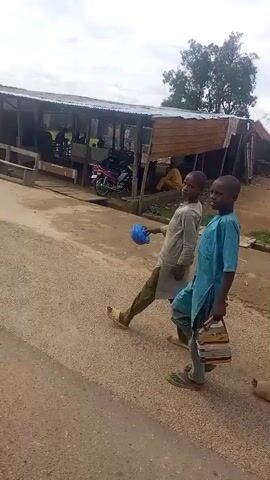 The video shows a street scene with several stalls and people. Two young men are walking in the foreground. One is wearing a grey robe and sandals, and the other is wearing a blue robe. Behind them, there are several makeshift stalls with corrugated iron roofs. One stall has motorcycles parked in front of it. Another stall appears to be selling goods, with bottles and containers displayed on tables. There are also benches and people sitting or standing around the stalls. The ground is a mix of dirt and gravel. The sky is overcast.