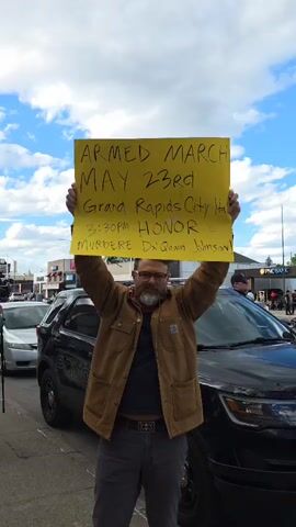 A man is standing in front of a black police car holding a yellow sign with black writing that reads "ARMED MARCH MAY 23rd Grand Rapids City Hall 3:30 PM HONOR MURDERED Da'Quan Johnson". The man has glasses, a beard, and is wearing a brown jacket. There are other people in the background, including one wearing a mask. The scene appears to be a protest or demonstration. The sky is visible and partly cloudy. A PNC Bank sign can be seen across the street.