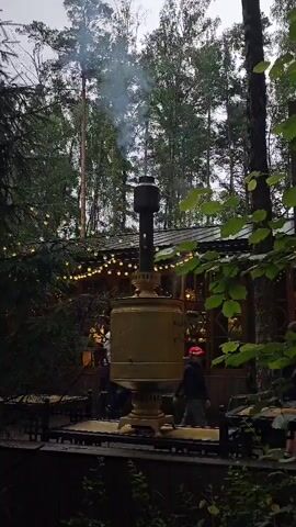 The video shows an outdoor scene with a large, ornate samovar in the center, emitting smoke from its chimney. The samovar appears to be made of brass or a similar metal and is placed on a raised platform. Behind the samovar, a rustic building with a dark roof and string lights is visible. The building seems to be a restaurant or cafe, as tables and chairs are set up in front of it. The surrounding environment is a forest, with tall trees and lush green foliage. A person walks past the samovar from right to left in the foreground. Another person walks into frame on the right side and appears to be interacting with something held in their hands. The lighting suggests it is either dusk or a cloudy day.