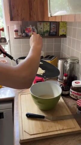 A person is making crepes. They are holding a ladle full of crepe batter over a hot pan, tilting the pan to spread the batter into a thin circle. The batter is pale yellow and appears to be a smooth liquid. The pan is dark and circular with a handle. In the foreground, a green bowl contains some of the crepe batter. In the background, there are kitchen appliances, including a toaster and a coffee maker, as well as shelves with boxes of tea. The kitchen appears to be well-lit.