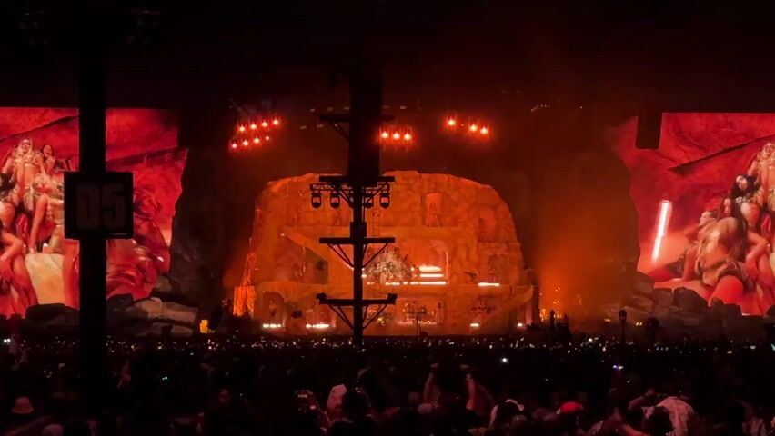 The video captures a wide shot of a large-scale live concert or music festival at night. A vast crowd of people fills the foreground, many holding up illuminated cell phones, creating a shimmering effect. An elaborate stage set in the background resembles a multi-tiered, glowing orange cave or rocky structure with various platforms and light fixtures. Two massive video screens flank the central stage. The left screen briefly displays a white "D5" marker on a dark background before showing women performers. The right screen continuously features close-up visuals of women, possibly dancers or the main artist, dressed in elaborate, sparkling costumes, dancing or posing against a vivid red, rocky backdrop. The stage is bathed in warm orange and red lights, with bright spotlights illuminating the set. Music, characterized by a rhythmic beat and what sounds like singing or chanting, is audible throughout. The overall atmosphere is dark, vibrant, and energetic, typical of a major musical performance.