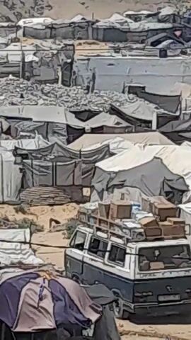 A white and blue van is parked on a dirt road. The van has a roof rack piled high with boxes. The background consists of numerous tents, suggesting a refugee camp or informal settlement. Some tents are white, while others are a muted tan color. In the foreground, a small market stall with various items is visible, partially obscured by a purple and grey umbrella. The overall impression is one of a transient or temporary living situation.