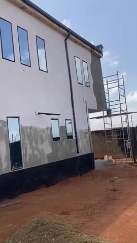The video showcases the exterior of a building under construction in Bar Gada, Nigeria. The building has a white facade with several tall, rectangular windows that reflect the blue sky and clouds, indicating it's daytime with fair weather. The lower half of the building's exterior wall is partially covered in grey plaster, suggesting ongoing renovation or construction work. A black foundation base runs along the bottom of the building.

In the foreground, piles of reddish-brown soil and gravel are visible, indicating a construction site. Wooden scaffolding or supports are present near the bottom of the frame. One section of the building features an air conditioning unit mounted on the wall, with a white pipe running upwards. Reflections in the windows offer glimpses of the surrounding environment, including what appears to be fencing and possibly other structures.

In one of the lower windows, two men are visible. One is sitting on something inside the building, and another is standing near the window. Their presence confirms human activity related to the construction or the building itself. The overall scene depicts a work in progress, typical of a developing area. The clear sky suggests a generally pleasant weather day, suitable for outdoor construction. There is no discernible emotional tone, as the video is purely observational. No significant text is visible in the video.