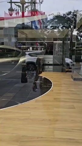 A silver sports car is displayed in a showroom. Reflections of a street scene, including buildings, traffic lights, and a man, are visible on the car's surface and the showroom window. The showroom appears to be a GR Garage, indicated by signage. The man, wearing a plaid shirt and shorts, is holding a smartphone and appears to be taking a photo or video. The showroom has wooden and tiled flooring.