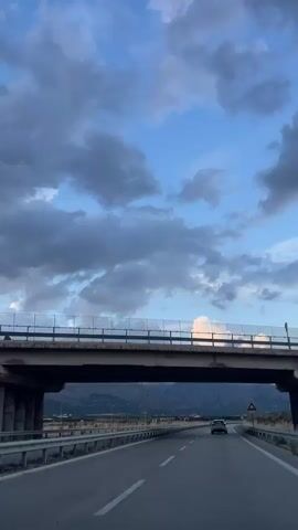 The video shows a view from a car driving on a highway. A bridge is visible overhead. The sky is blue with clouds. Mountains are in the distance. One car is ahead on the highway.