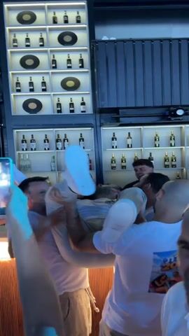 The video captures a group of young men in what appears to be a bar or restaurant. In the foreground, three men are hoisting another man in the air, supporting him by his legs and torso. The man being lifted is wearing a white t-shirt and light-colored pants, with white sneakers. He is in a horizontal position, with his feet pointing upwards. The men holding him are also wearing white t-shirts. The background reveals shelves stocked with bottles, presumably alcoholic beverages, and spotlights illuminating the area. The lighting shifts between warm yellow and cool blue hues, suggesting dynamic lighting effects common in entertainment venues. The atmosphere seems celebratory, with the individuals appearing to be enjoying themselves.