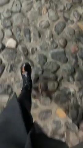 The video starts with a close-up of a stone path, then pans up to show a woman walking along the path at night. She has long brown hair and is wearing a green hooded jacket and a tan-colored bag. The path is lined with a black metal fence and a building with a Coca-Cola sign. There is a large metal box structure in front of the building.