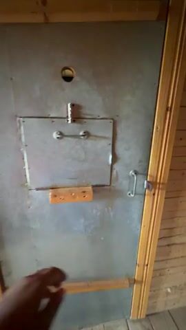 A person's hand in a red sleeve reaches for a hook latch on a door. The door opens to reveal a dimly lit, rustic cell with stone walls and a wooden floor and ceiling. A small, barred window is visible on the far wall, casting a faint orange glow into the room. Two metal bunks, suspended by chains, are attached to the wall.