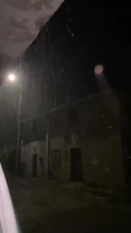The video begins with a dark, nighttime scene of a rural or suburban street. A street light illuminates some of the area, casting a soft glow on a two-story building with a dark facade. Rain is visible falling, catching the light in streaks. The camera pans to the right, revealing more of the building and a dark, open doorway. The scene is low-light and somewhat grainy, with the rain creating a dynamic element. There are no people visible in the video, and the focus is on the building and the atmospheric conditions. The video ends with a continuation of the rain and the dark surroundings.