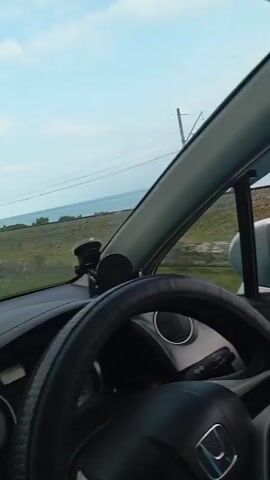 The video, shot from inside a car, shows a person's hand resting on the car door. Outside the window, a railroad track runs parallel to the road, and beyond that, a body of water, likely the sea, is visible under a cloudy sky. The car appears to be moving, indicated by the motion blur of the landscape and the reflection in the side mirror. The overall impression is of a scenic drive along a coastal route.