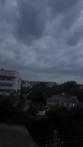 The video shows a view of a residential area from an elevated position. The sky is overcast with dark clouds. Several buildings, including apartment buildings and houses, are visible amidst trees. Some windows in the houses are illuminated, suggesting it is evening or nighttime. The foreground shows a dark railing or ledge. The overall atmosphere is gloomy.