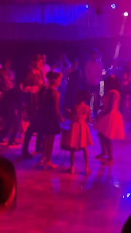 The video shows a stage performance with a group of people. In the foreground, two young girls in dresses are standing near a drum. One girl, on the left, is facing away from the camera and appears to be interacting with the drum. The other girl, in the center, is also facing away from the camera and is holding drumsticks. To the left of these two girls, a person in dark clothing is looking towards them. In the background, several other people are visible, some of whom are clapping. The stage is lit with vibrant pink and blue lights, with smoke creating a hazy atmosphere. There are also some stage lights visible, including spotlights. The overall impression is of a lively performance with music and dancing.